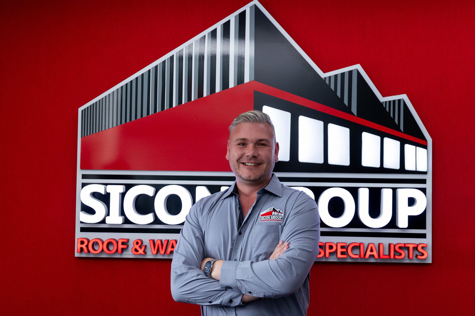 Jurgens Barnard stands confidently with arms folded in front of the iconic red Sicon Group wall and company logo, wearing a grey button-up shirt.