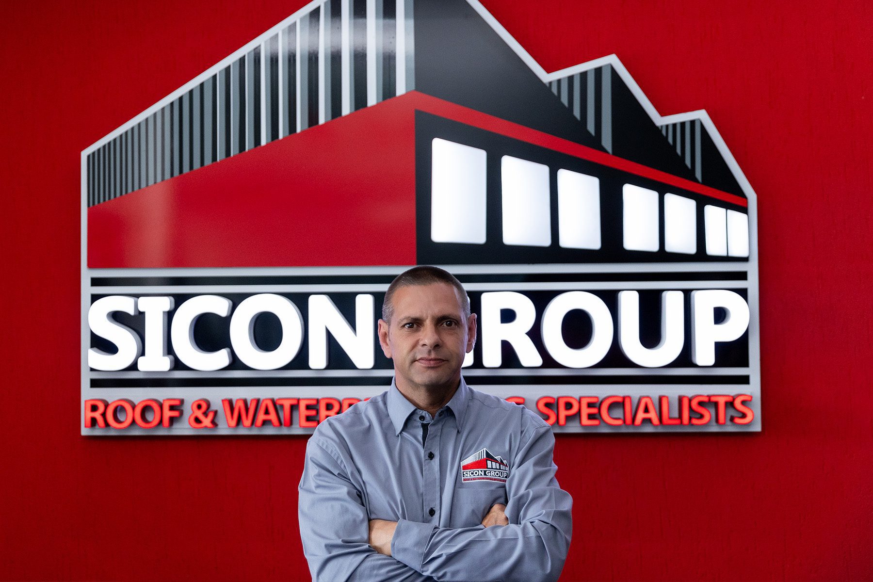 Portrait of Sean Souries, Sales Executive at Sicon Group, standing confidently in front of the company’s red and black logo wall.